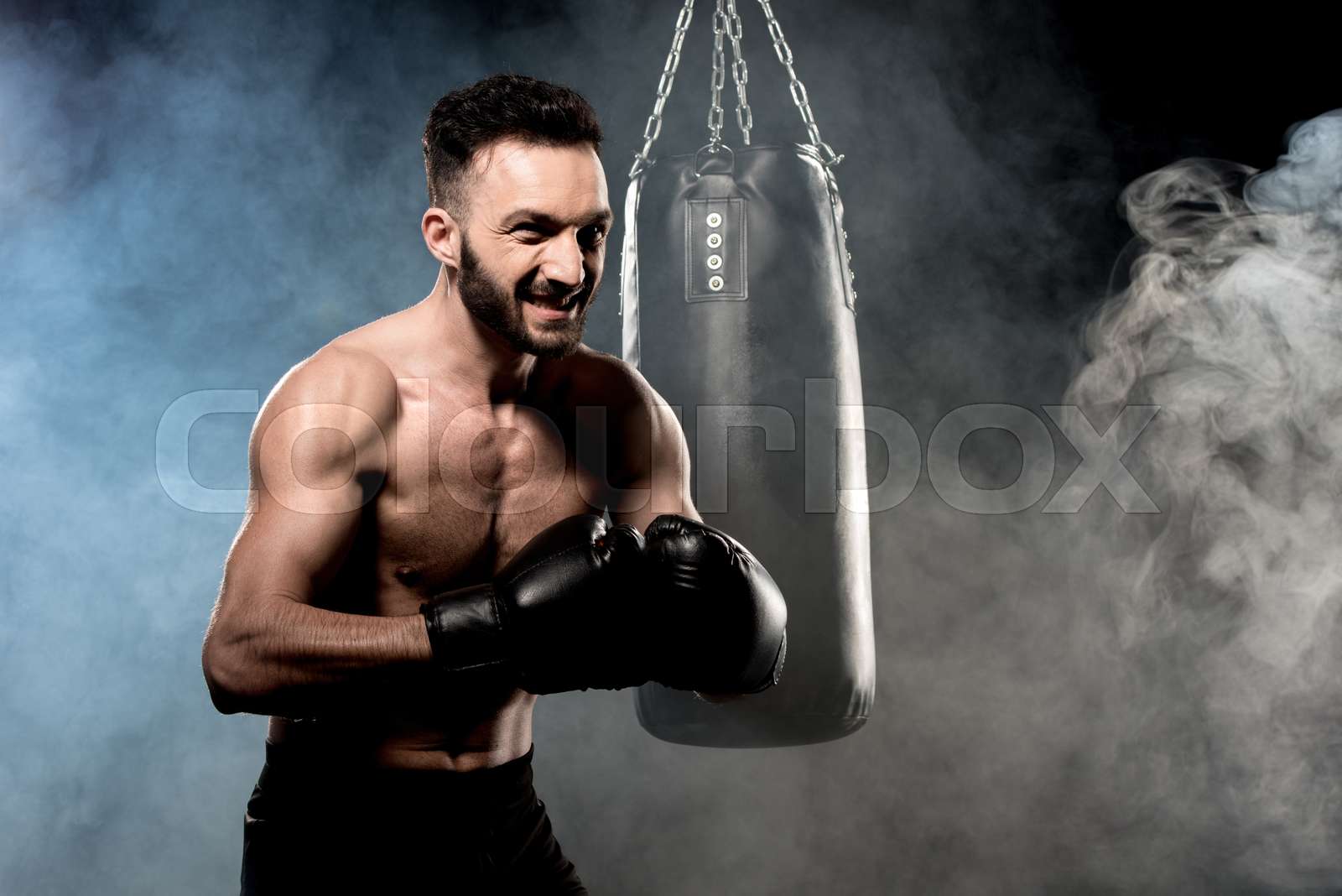 angry boxer standing in boxing pose near punching bag on black with