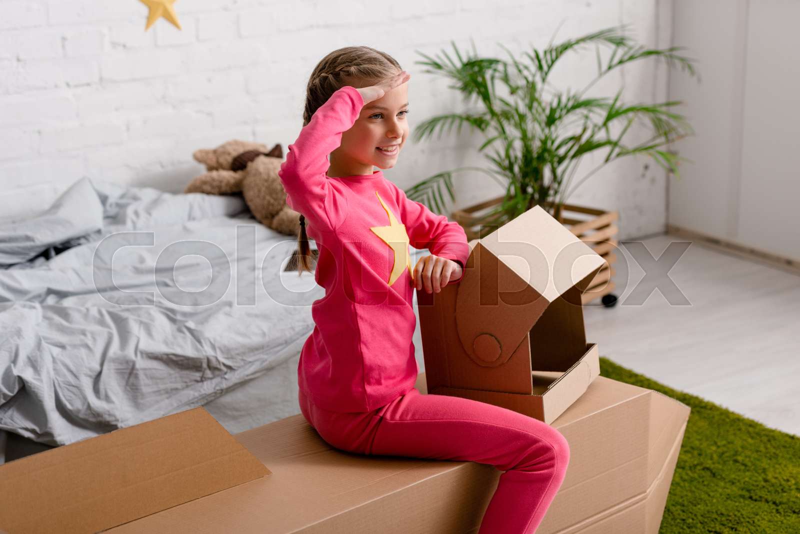 Smiling kid with helmet saluting while sitting on cardboard rocket in
