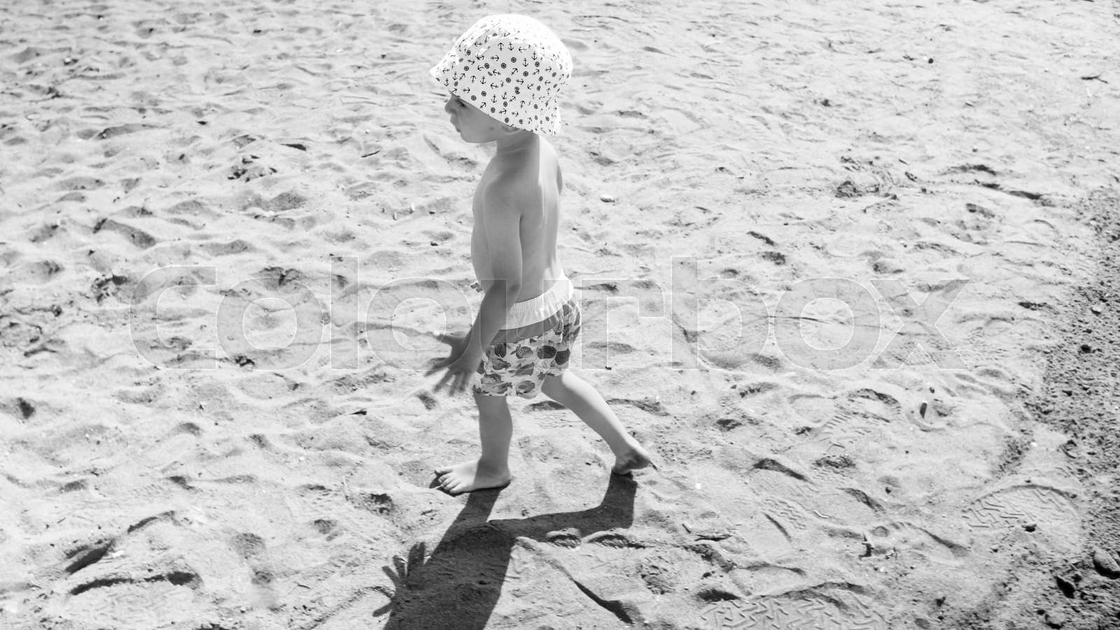 Black and white image of little toddler boy walking on hot sand at sea