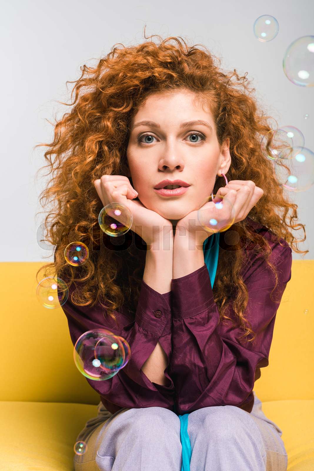 attractive curly redhead woman looking at camera and touching face near soap bubbles on white