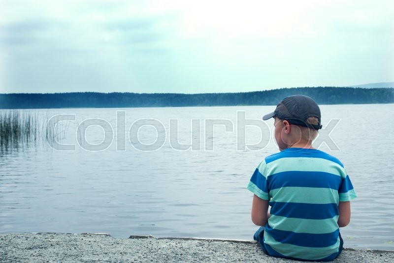 sad child sitting alone at the lake on a cloudy day back view