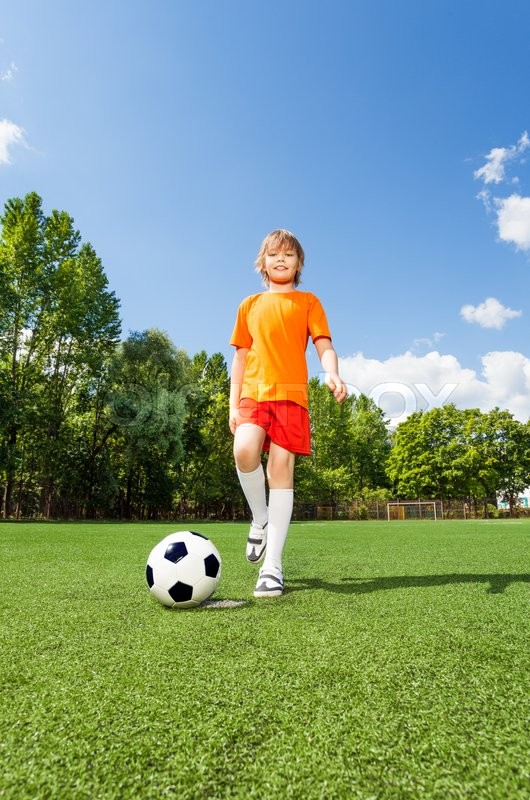 happy boy kicking football with one leg on green grass of field in
